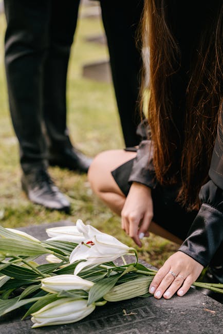 A person dressed in dark clothing kneels beside a grave, arranging a floral display consisting of white lilies with long green leaves. The lilies are freshly cut, with smooth, slightly curved petals a